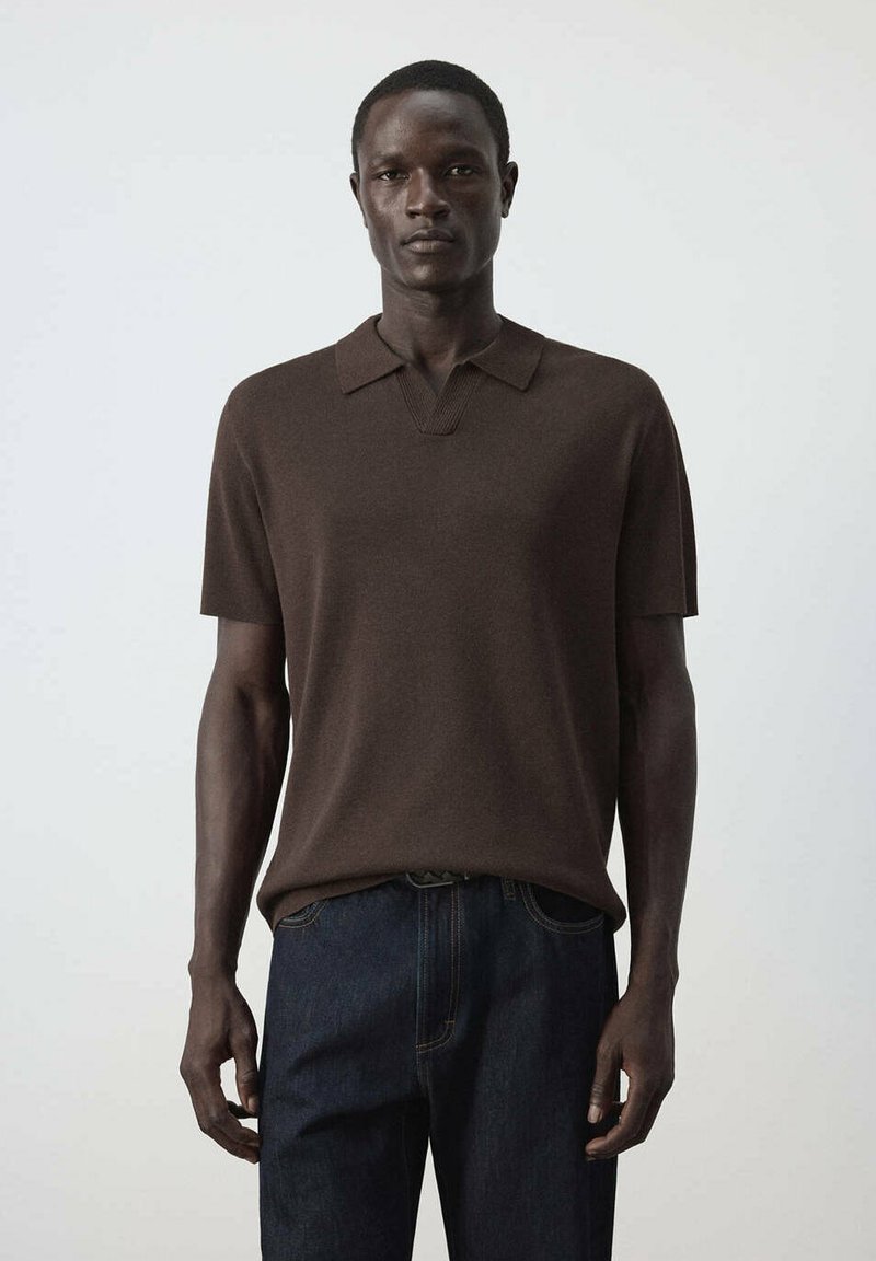 Homme debout face à l'avant, portant une chemise à manches courtes marron foncé avec col et un jean bleu foncé sur un fond clair uni.