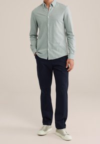 Světlounce zelená košile se zapínáním na knoflíky a dlouhými rukávy, vyrobená z měkké látky, v kombinaci s námořnicky modrými kalhotami a světlými teniskami.