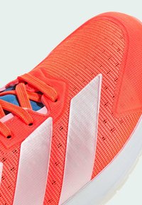 Svěží korálová sportovní bota s bílými pruhy, síťovinovým svrškem a odpovídajícími korálovými tkaničkami na světlém pozadí.