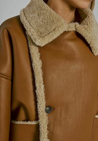 Vrouw in een bruine leren jas met beige schapenvacht trim rond de kraag en randen, gesloten met een zichtbare knop.