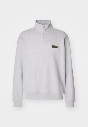 Sudadera de color gris claro con media cremallera en el cuello, puños y dobladillo acanalados, que presenta un logo de cocodrilo bordado en verde en el pecho.
