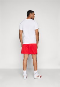 White short-sleeve t-shirt, red mesh athletic shorts, white socks with logo, red and white sneakers, standing with back facing camera.