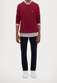 Röd kabelstickad tröja med rund halsringning, lager-på-lager över en vit och blå randig skjorta, kombinerad med mörka byxor och vita sneakers.