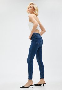 Mujer con pelo corto rubio que lleva una blusa blanca sin tirantes, jeans ajustados de color azul oscuro y zapatos de tacón bajo negros, de pie de perfil.
