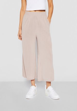 Persoon die beige wijde cropped broek met elastische tailleband draagt, witte cropped tanktop en witte sneakers op een grijze vloer.