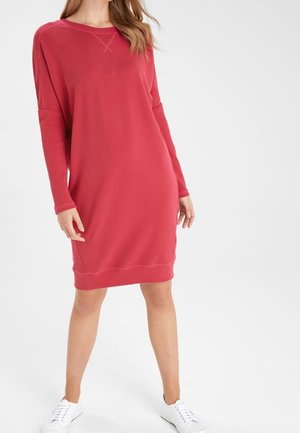 Femme portant une robe rouge ample à manches longues et des baskets blanches, debout sur un fond blanc uni.