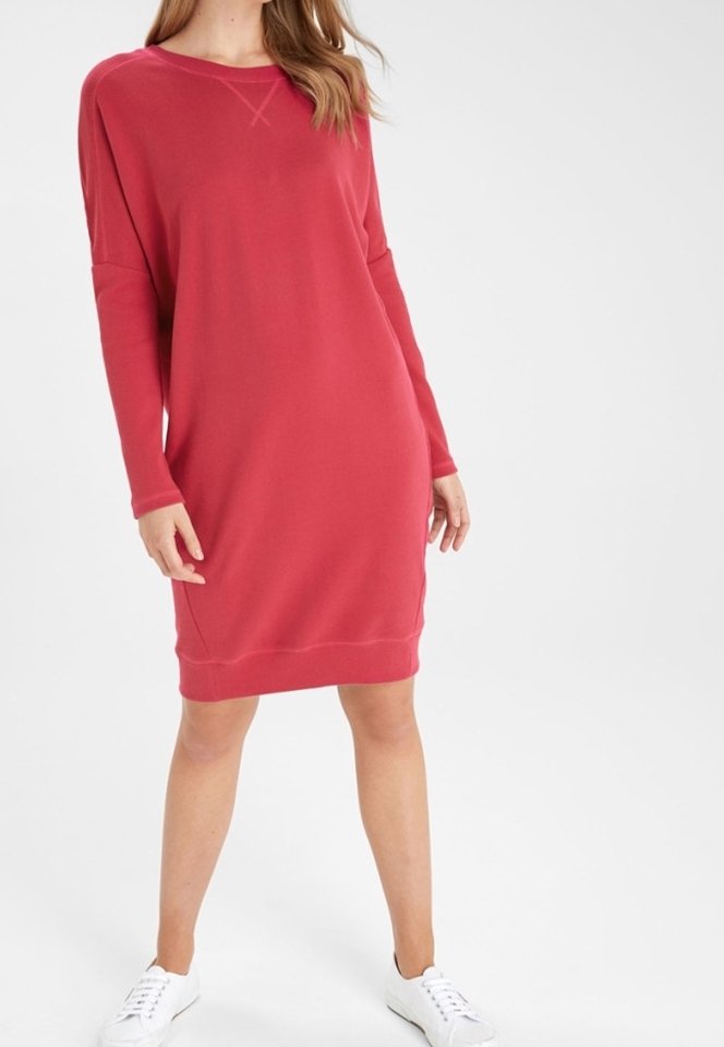 Femme portant une robe rouge ample à manches longues et des baskets blanches, debout sur un fond blanc uni.