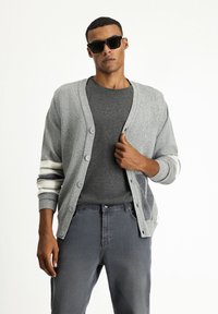 Cárdigan gris con cuello en V, botones en el frente y rayas blancas en contraste en las mangas. Lleva puesto una camiseta gris oscuro y jeans grises.