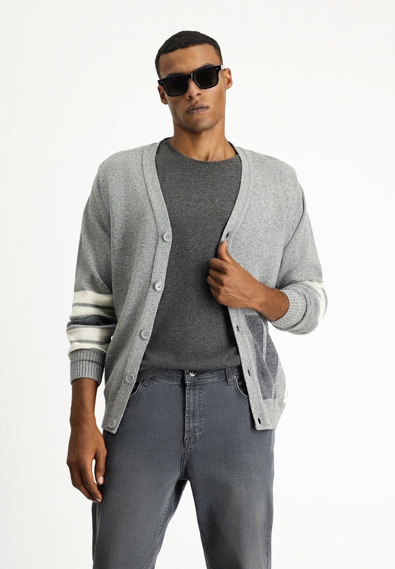 Cárdigan gris con cuello en V, botones en el frente y rayas blancas en contraste en las mangas. Lleva puesto una camiseta gris oscuro y jeans grises.