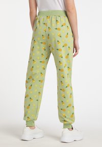 Grüne Sweatpants mit einem wiederholten Muster aus Ananas und Zitronen, elastischem Bund und Bündchen. Von hinten mit weißen Turnschuhen gezeigt.
