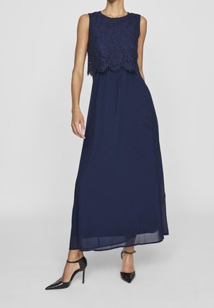 Frau in einem ärmellosen, marineblauen Maxikleid mit Spitzenoberteil und schwarzen High Heels mit Knöchelriemen, stehend vor einem schlichten Hintergrund.