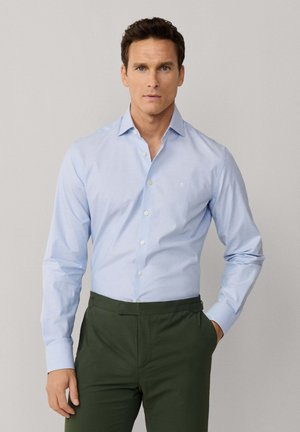 Man die een lichtblauw overhemd met knopen draagt, ingestopt in donkergroene broek, staand met één hand in de zak tegen een neutrale achtergrond.