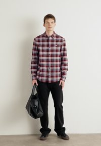 Camicia a quadri rossa e blu con colletto a bottoni, abbinata a pantaloni neri. Una borsa a tracolla grigia è tenuta in una mano. Tessuto morbido, design casual.