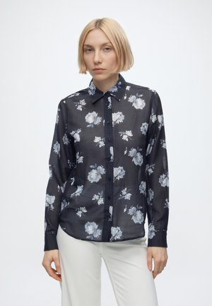 Junge Frau mit kurzen blonden Haaren, die eine transparente schwarze Bluse mit langen Ärmeln und weißen Blumenmustern sowie weiße Hosen vor einem schlichten Hintergrund trägt.