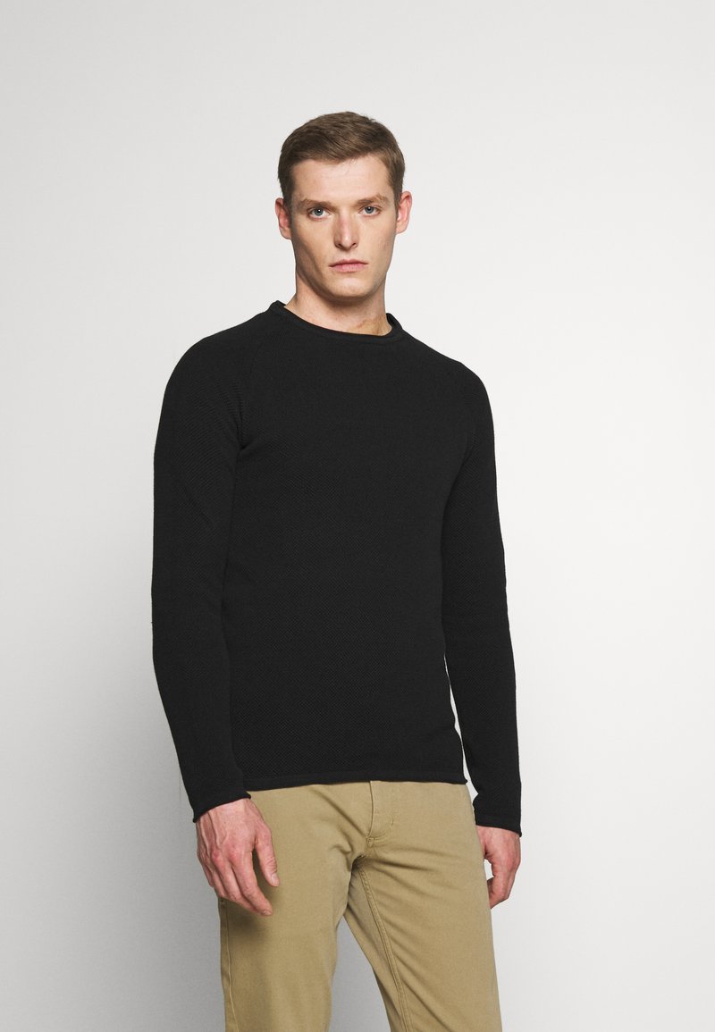 Schwarzer Pullover mit langen Ärmeln, einem strukturierten Strickmuster, Rundhalsausschnitt und tailliertem Schnitt, kombiniert mit beigen Hosen.