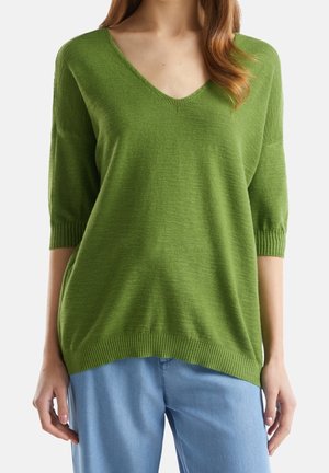 Kvinde iført en løs grøn V-hals sweater med albuelange ærmer og lyseblå bukser, stående med arme afslappet langs siden.
