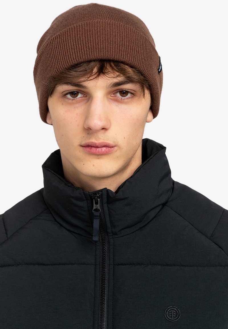 Bruine geribbelde gebreide beanie gedragen bij een zwarte gewatteerde jas met een hoge kraag en ritssluiting, wat de minimale uitstraling benadrukt.