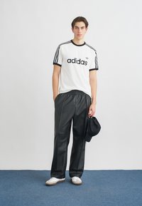 Biele tričko s čiernymi pruhmi a logom "adidas", v kombinácii s čiernymi saténovými teplákmi. Doplnky zahŕňajú čiernu šiltovku a biele topánky.