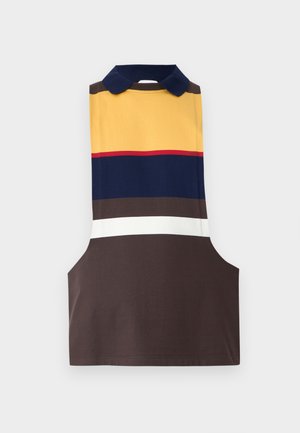 Bărbat stând sprijinit de un perete simplu, purtând un tricou polo fără mâneci cu dungi, pantaloni albastru aprins, pantofi negri și un colier cu pandantiv.