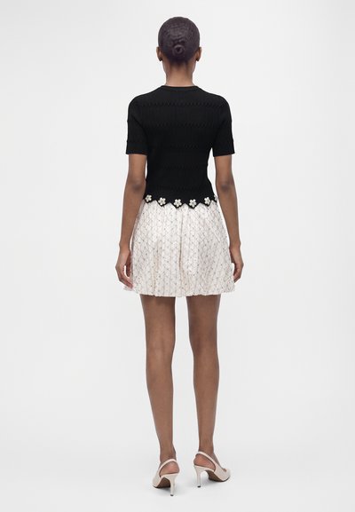 Femme debout, vue de dos, portant un haut noir à manches courtes avec bordure fleurie, une jupe blanche à motifs jusqu’aux genoux et des talons blancs.