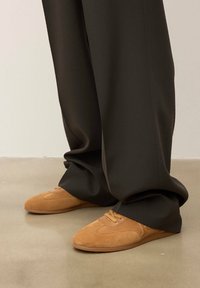 Sarkanā zamšāda apavi ar apaļu purngali, ar šuvēm un auklām, kopā ar tumšiem, platajām biksēm. Grīdas segums ir neitrāls.