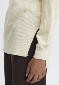 Stickad tröja i ljusbeige, med ribbat mönster, sidoflikar och en avslappnad passform. Matchas med mörkbruna byxor.