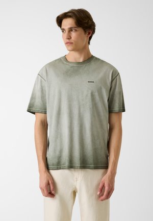 Młody mężczyzna w wyblakłej zielonej koszulce Guess i jasnobeżowych spodniach, patrzący w lewo na tle jednolitej białej ściany.