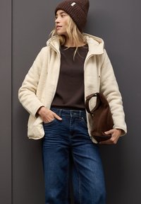 Chaqueta de forro polar crema con capucha, suéter marrón, jeans de denim azul y bolso de ante marrón. La modelo lleva un gorro de punto marrón oscuro.