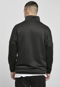 Sweatshirt noir zippé avec col haut, en tissu lisse. Présente des manches longues et amples ainsi qu'une coupe décontractée. Pas de logos visibles.
