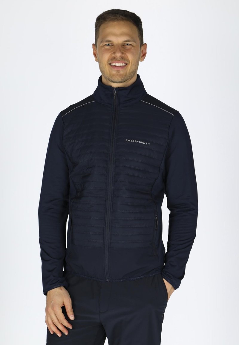 Marineblå zip-up jakke med polstret front, glatte ærmer, reflekterende detaljer og sidelommer for funktionalitet. Letvægts og tætsiddende design.