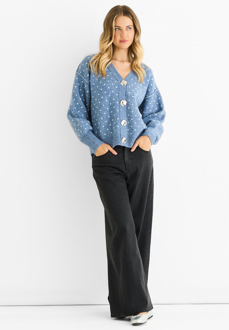 Woman standing with hands in pockets wearing blue polka-dot buttoned cardigan, black wide-leg jeans, and silver flats on white background.