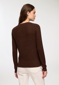 Maglione marrone lavorato a maglia con orlo e polsini a coste, caratterizzato da un scollo rotondo e una superficie testurizzata. Il modello indossa pantaloni chiari.