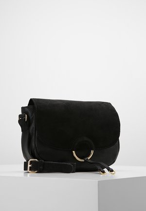 Black leather crossbody bag with suede flap, gold ring detail, and adjustable strap placed on white surface against light gray background.