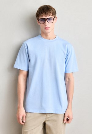 Tricou scurt, de culoare albastru deschis, cu decolteu rotund, dintr-un material neted. Purtați cu pantaloni bej și ochelari mari cu tentă.