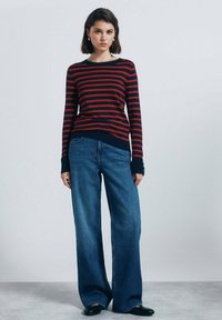 Rood en marineblauw gestreepte gebreide trui met lange mouwen, gecombineerd met brede blauwe denim jeans. Zwarte schoenen en minimale accessoires.