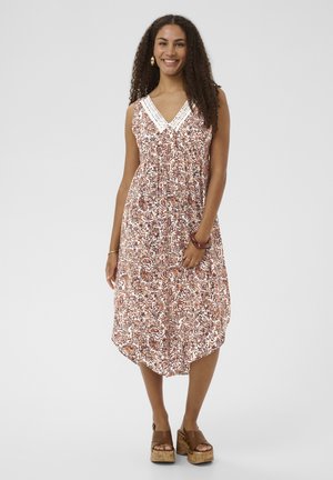 Kvinde med krøllet hår, iført en ærmeløs, mønstret midikjole med V-hals og platformsandaler, smilende mod en ensfarvet baggrund.