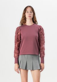 Maglione color lampone con dettagli a volant texturizzati sulle maniche trasparenti, abbinato a una gonna plissettata grigia. Collo tondo semplice.