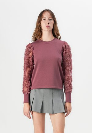 Sweatshirt couleur framboise avec des volants texturés sur des manches transparentes, associé à une jupe plissée grise. Encolure ronde simple.
