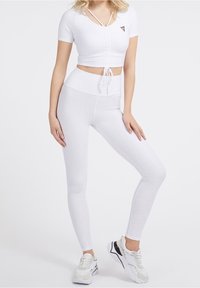 Žena v bílých texturovaných legínách s vysokým pasem a odpovídajícím krátkým rukávem crop topem se stahovací šňůrkou, doplněná bílými a černými robustními teniskami.