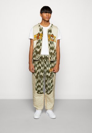 Kamuflasjemønstret quilted vest og bukser i grønn og beige, med oransje "MARKET"-bokstaver, hvit t-skjorte og hvite joggesko.