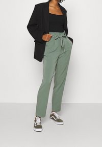 Blazer noir sur un débardeur noir ; pantalon taille haute vert clair avec une ceinture à nouer ; baskets vert olive et blanches.