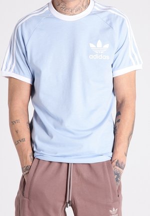 Personne portant un t-shirt Adidas bleu clair avec des rayures blanches et un logo, associé à un pantalon de survêtement Adidas mauve avec des poches latérales.