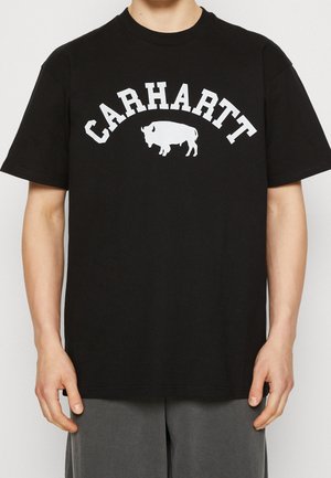 Personne portant un t-shirt Carhartt noir avec un logo de buffle blanc centré sous le nom de la marque en lettres majuscules grasses.