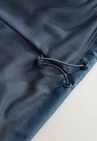Tissu bleu foncé avec de petites perforations, doté d'un cordon de serrage passé à travers deux œillets en métal avec un réglage par boutonnet en plastique.