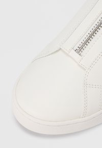 Λευκό δερμάτινο αθλητικό παπούτσι με στρογγυλεμένη μύτη, διακοσμημένο με ραφές και πλαϊνό μεταλλικό φερμουάρ. Η σόλα με απαλή υφή βελτιώνει την πρόσφυση.