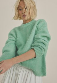 Mint green, fuzzy knit sweater with a high neckline and ribbed cuffs. Soft texture and relaxed fit. Model's arms are partially outstretched.