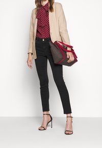Mujer vistiendo un blazer beige, blusa roja con estampado y lazo, jeans ajustados negros, sandalias de tacón alto negras, sosteniendo un bolso marrón con asas rojas.