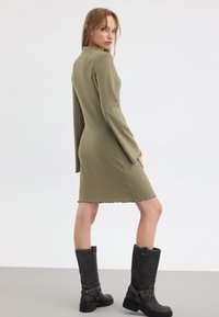 Olivengrünes, strukturiertes knielanges Kleid mit langen Ärmeln, kombiniert mit schwarzen kniehohen Stiefeln, die einen Schnallenverschluss und silberne Hardware-Details aufweisen.