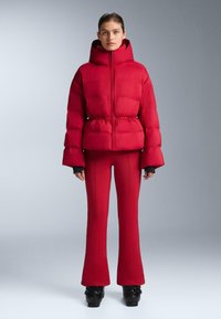 Piros bélelt dzseki magas nyakkal és elasztikus derékrésszel, hozzá illő, testhez simuló piros szárabban szűkülő nadrággal, fekete lábbelivel kiegészítve. Sima textúra.