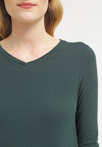 Długos LEWY, dekolt w serek, koszula wykonana z miękkiej, ciemnozielonej tkaniny o gładkiej fakturze i lekkim rozciągliwości, z dekoracyjnym przeszyciem wzdłuż dekoltu.